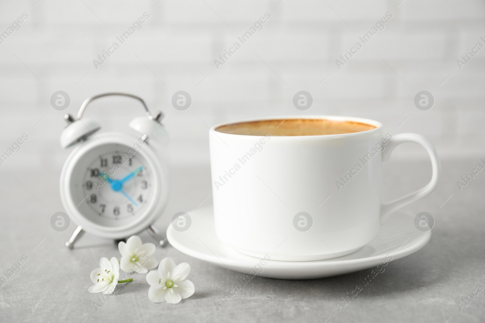 Photo of Cup of morning coffee, flowers and alarm clock on light grey table, closeup