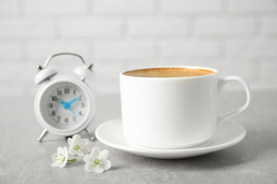 Cup of morning coffee, flowers and alarm clock on light grey table, closeup