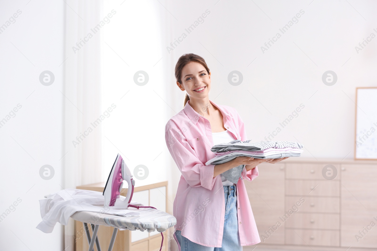 Photo of Young pretty woman holding stack of clean laundry at ironing board indoors