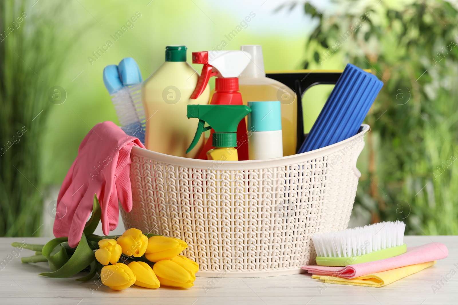 Photo of Spring cleaning. Plastic basket with detergents, supplies and beautiful flowers on white wooden table outdoors