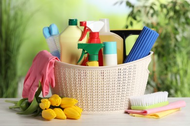 Photo of Spring cleaning. Plastic basket with detergents, supplies and beautiful flowers on white wooden table outdoors