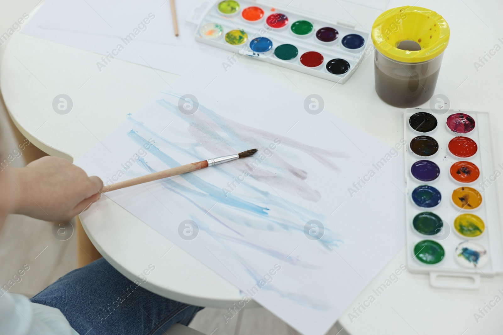 Photo of Little girl painting with brush and watercolor at white table, closeup
