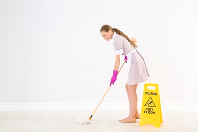 Photo of Young chambermaid with mop near WET FLOOR sign indoors