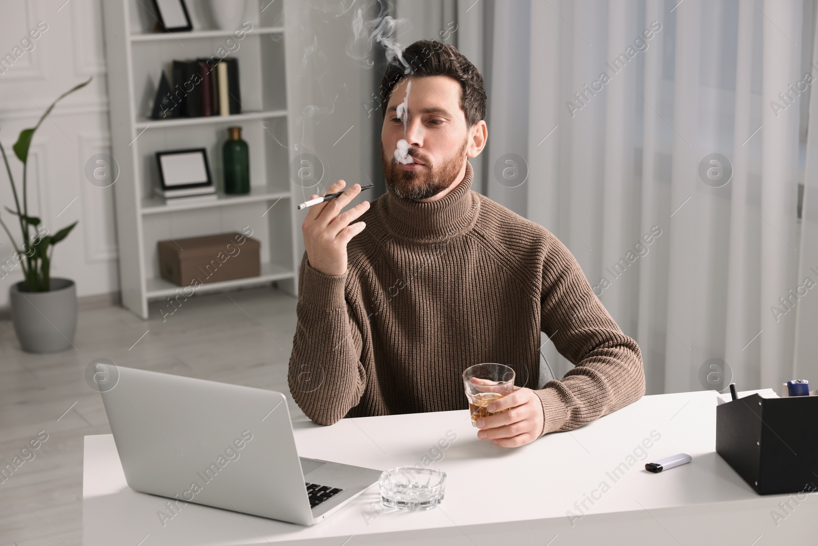 Photo of Man using long cigarette holder for smoking and holding glass of whiskey at workplace in office