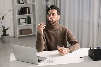 Photo of Man using long cigarette holder for smoking and holding glass of whiskey at workplace in office