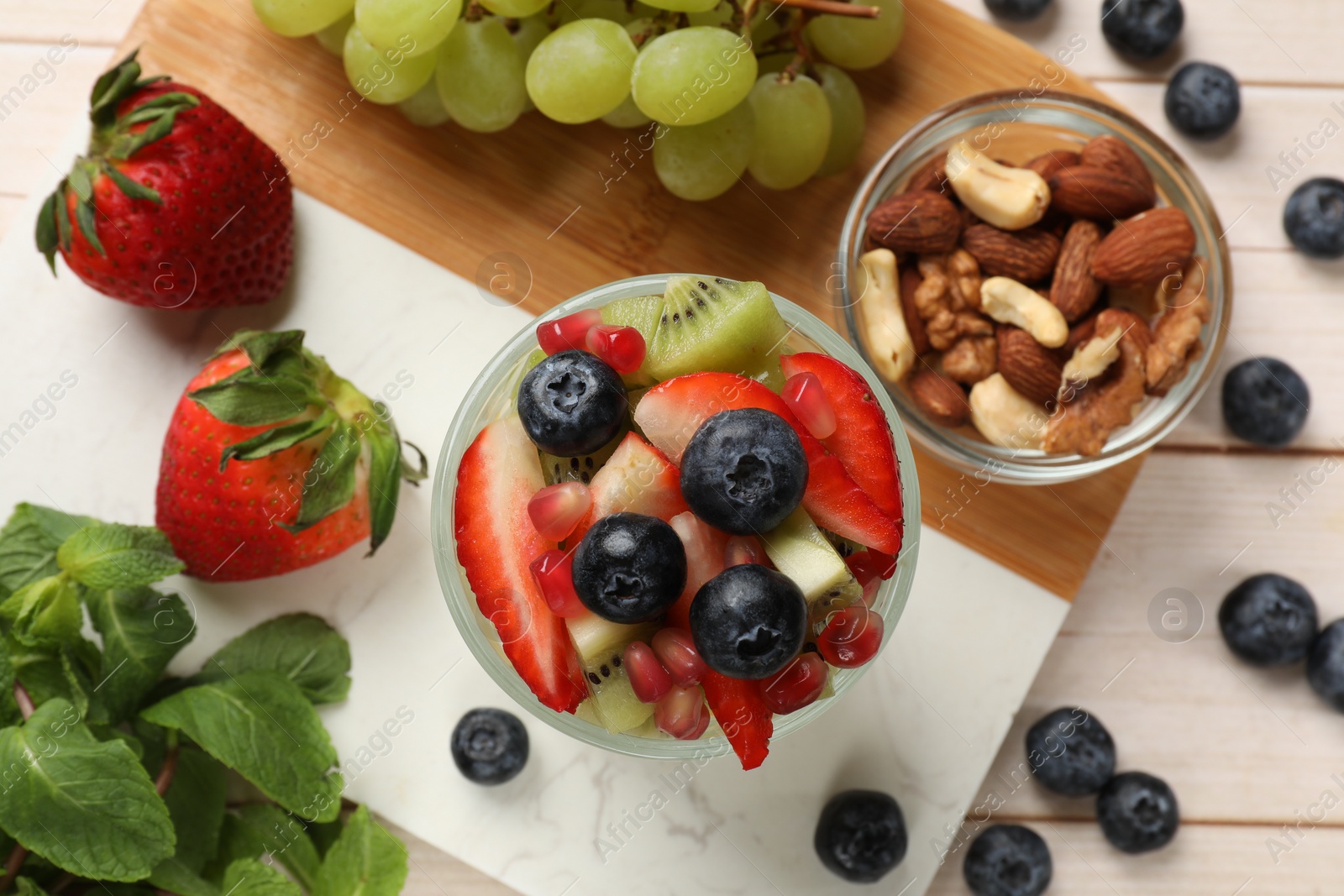 Photo of Healthy breakfast. Delicious fruit salad in glass, nuts and ingredients on light wooden table, flat lay