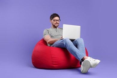Photo of Happy man with laptop sitting in beanbag chair against lilac background