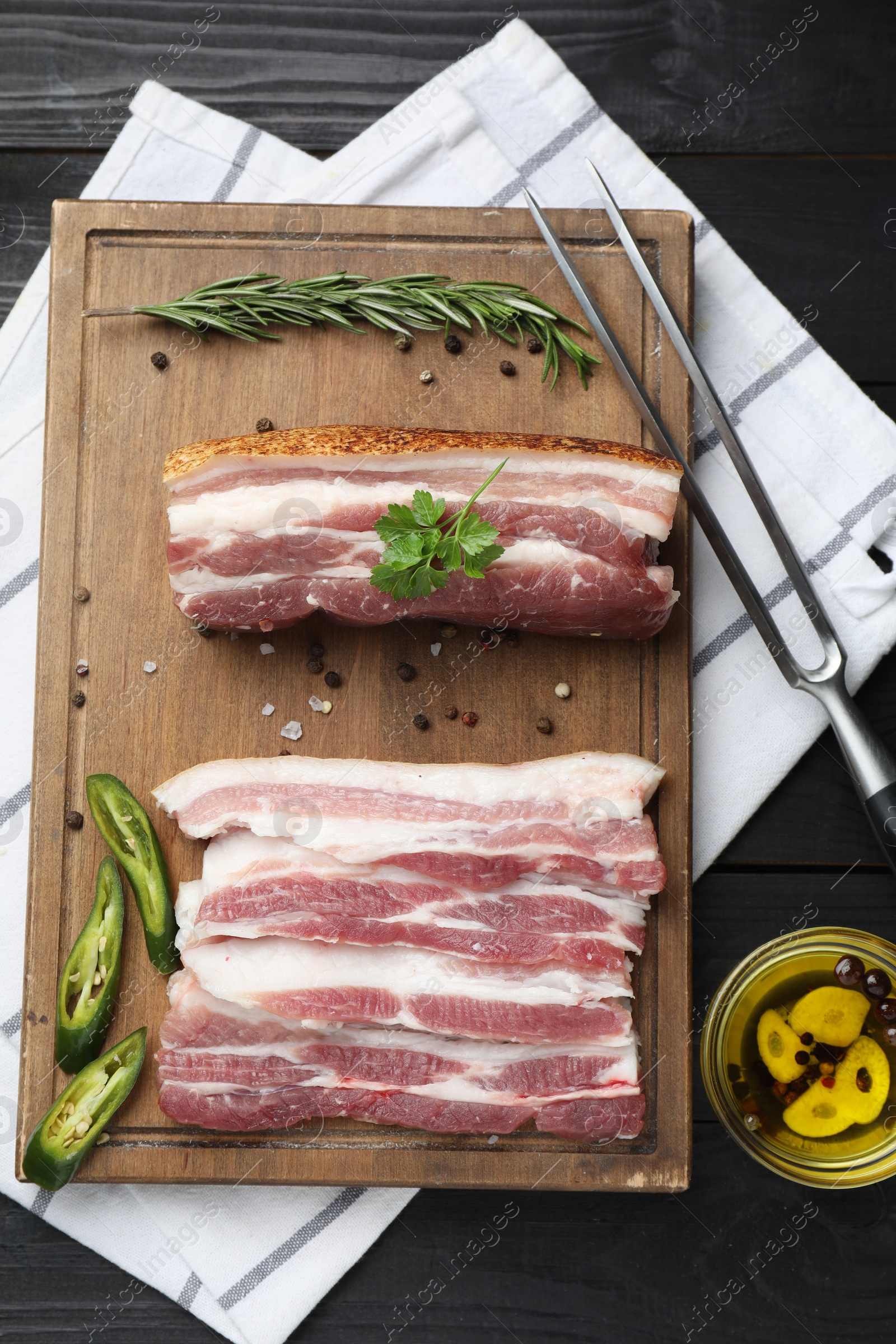 Photo of Pieces of raw pork belly, chili pepper, peppercorns, rosemary and parsley on black wooden table, flat lay