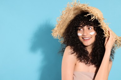 Beautiful young woman in straw hat with sun protection cream on her face against light blue background, space for text