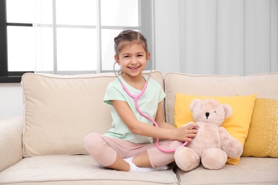 Photo of Cute child playing doctor with stuffed toy on sofa in hospital