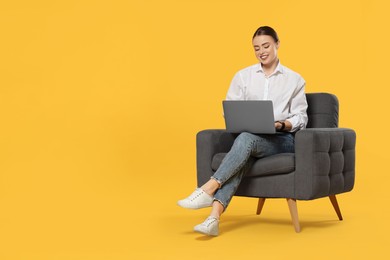 Happy woman with laptop sitting in armchair on orange background. Space for text