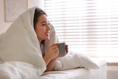 Photo of Beautiful young woman covered with warm blanket enjoying hot drink on bed at home. Space for text