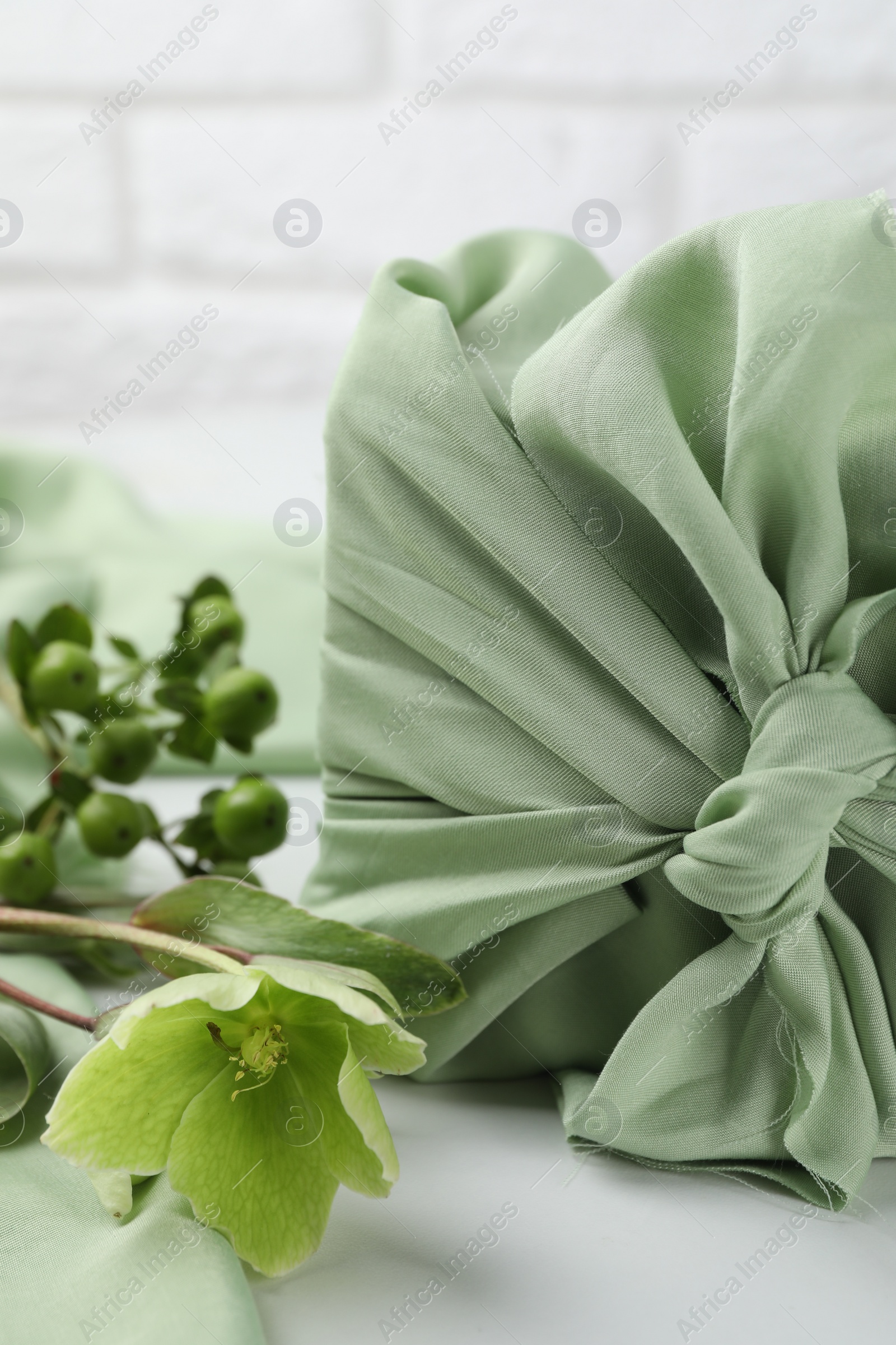 Photo of Furoshiki technique. Gift packed in green fabric and plants for decor on white table, closeup