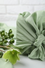 Photo of Furoshiki technique. Gift packed in green fabric and plants for decor on white table, closeup