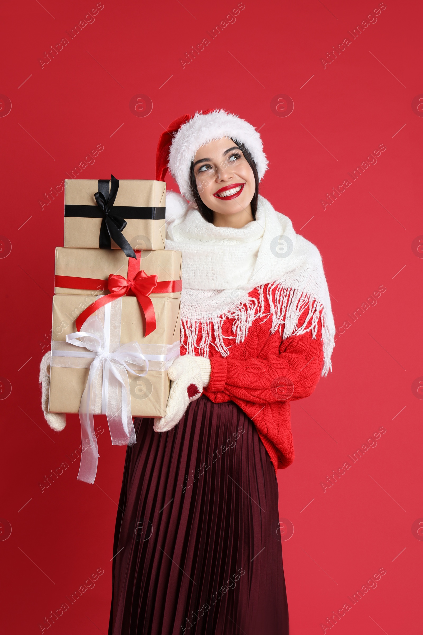 Photo of Woman in Santa hat, scarf and sweater holding Christmas gifts on red background