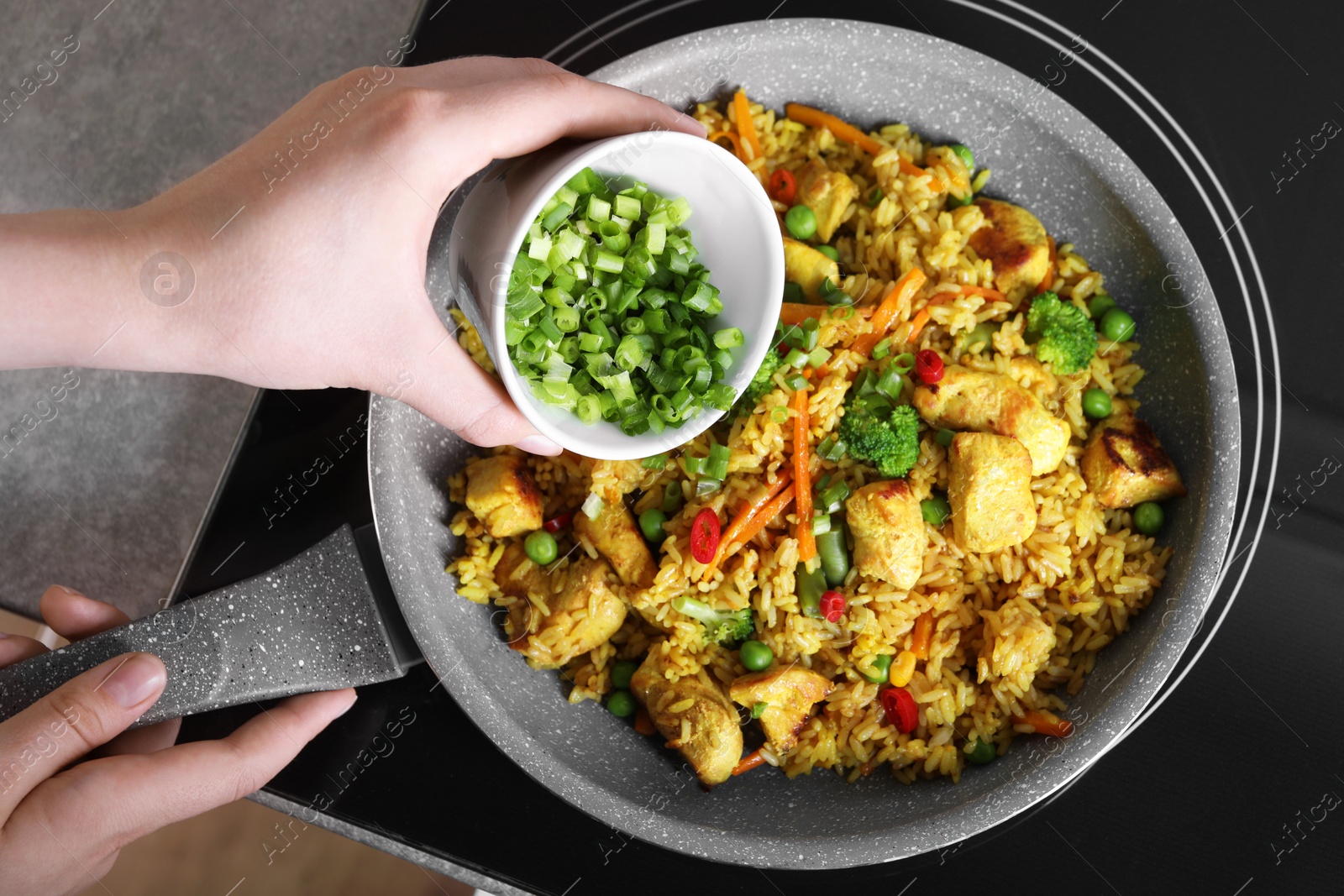 Photo of Woman adding cut green onion to rice with meat and vegetables in frying pan, top view