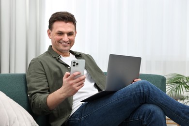 Photo of Happy man with laptop using smartphone on sofa at home
