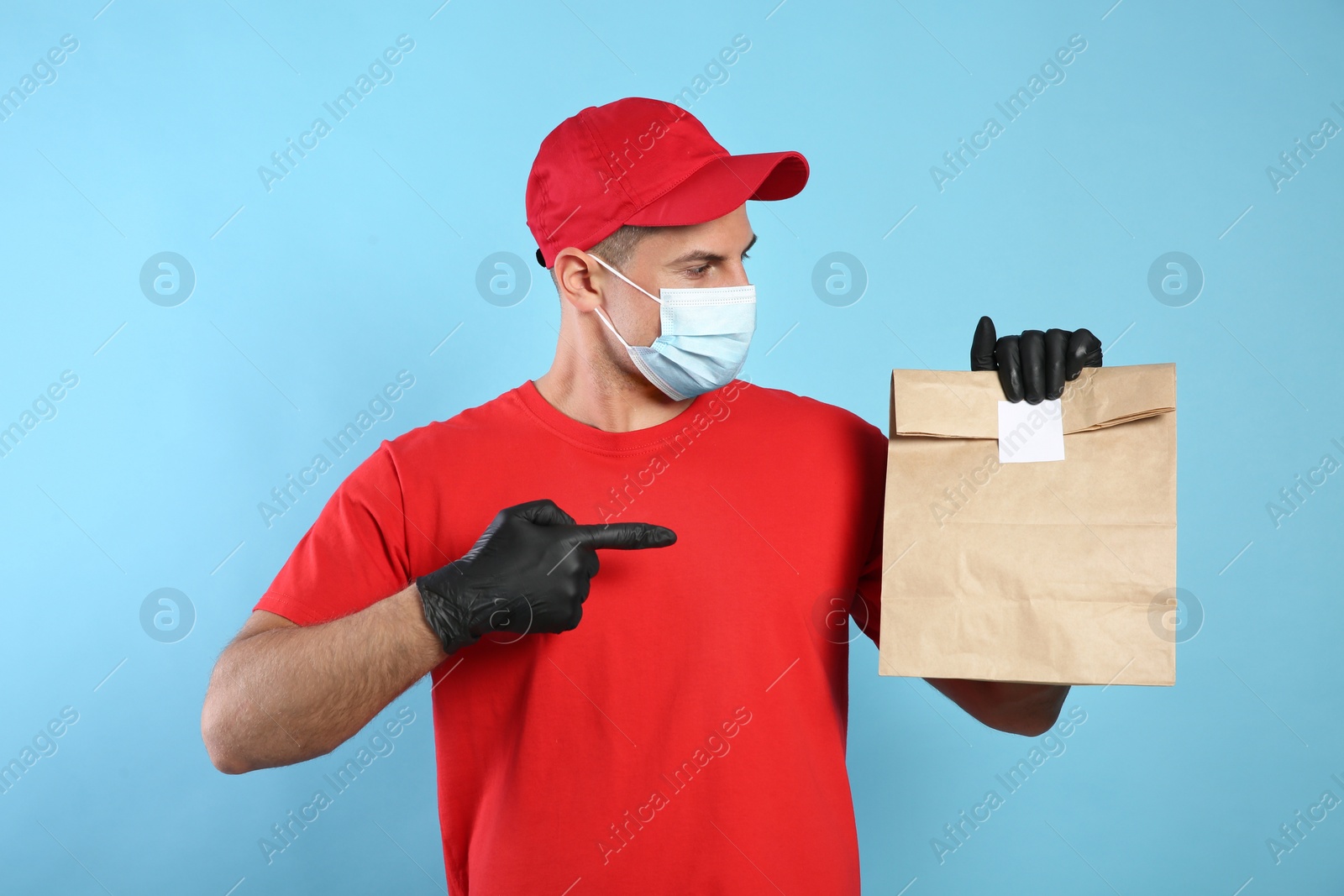 Photo of Courier in medical mask holding paper bag with takeaway food on light blue background. Delivery service during quarantine due to Covid-19 outbreak