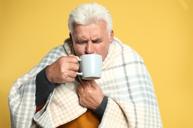 Senior man wrapped in blanket drinking hot beverage on yellow background. Cold symptoms