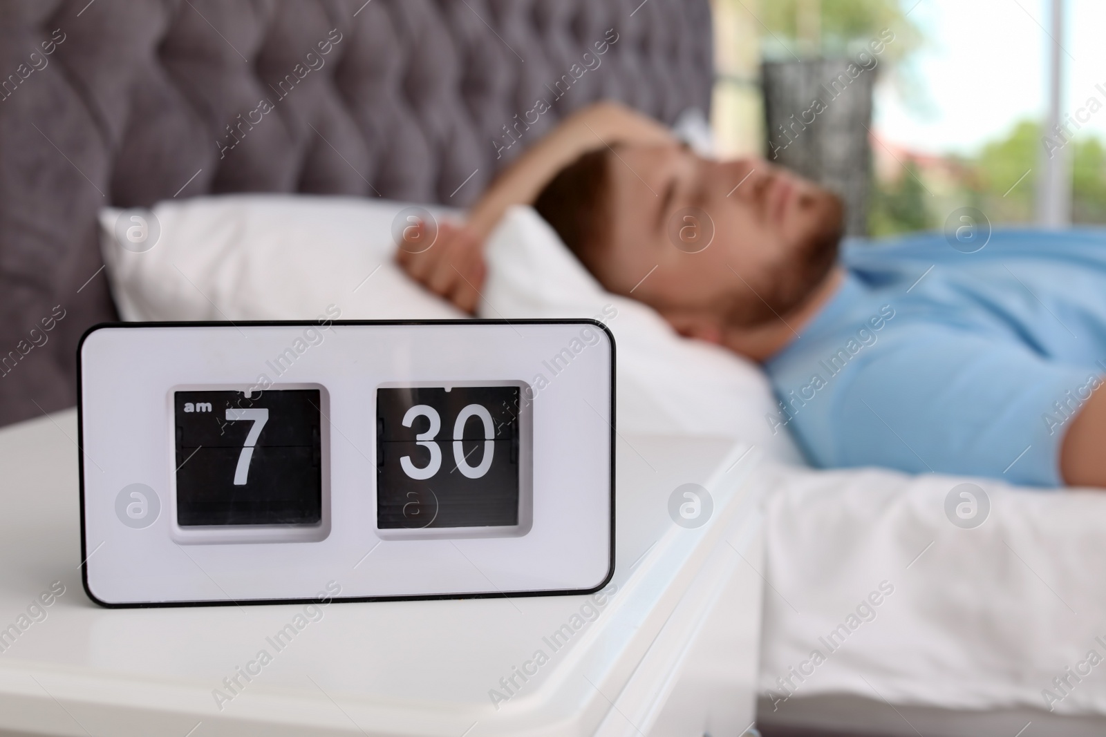 Photo of Alarm clock on table and young man sleeping in bed at home