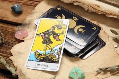 Photo of The Fool near other tarot cards with gemstones and old book on wooden table, closeup