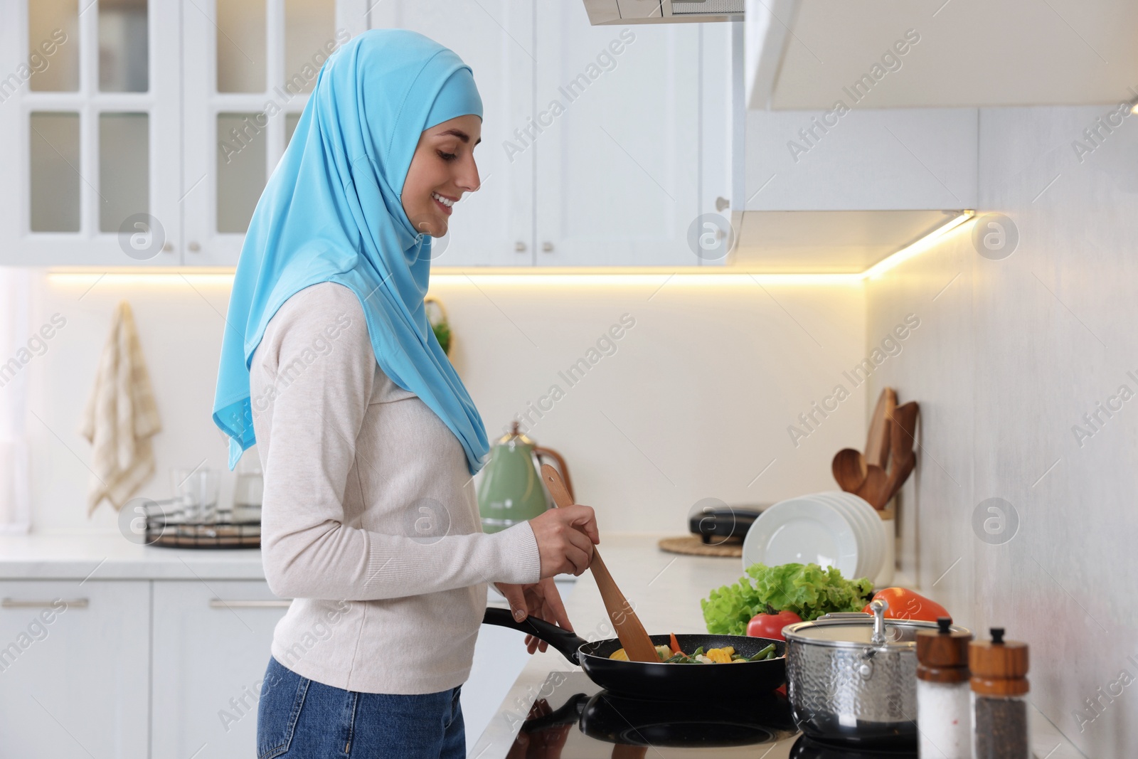 Photo of Muslim woman cooking dish in frying pan on cooktop indoors