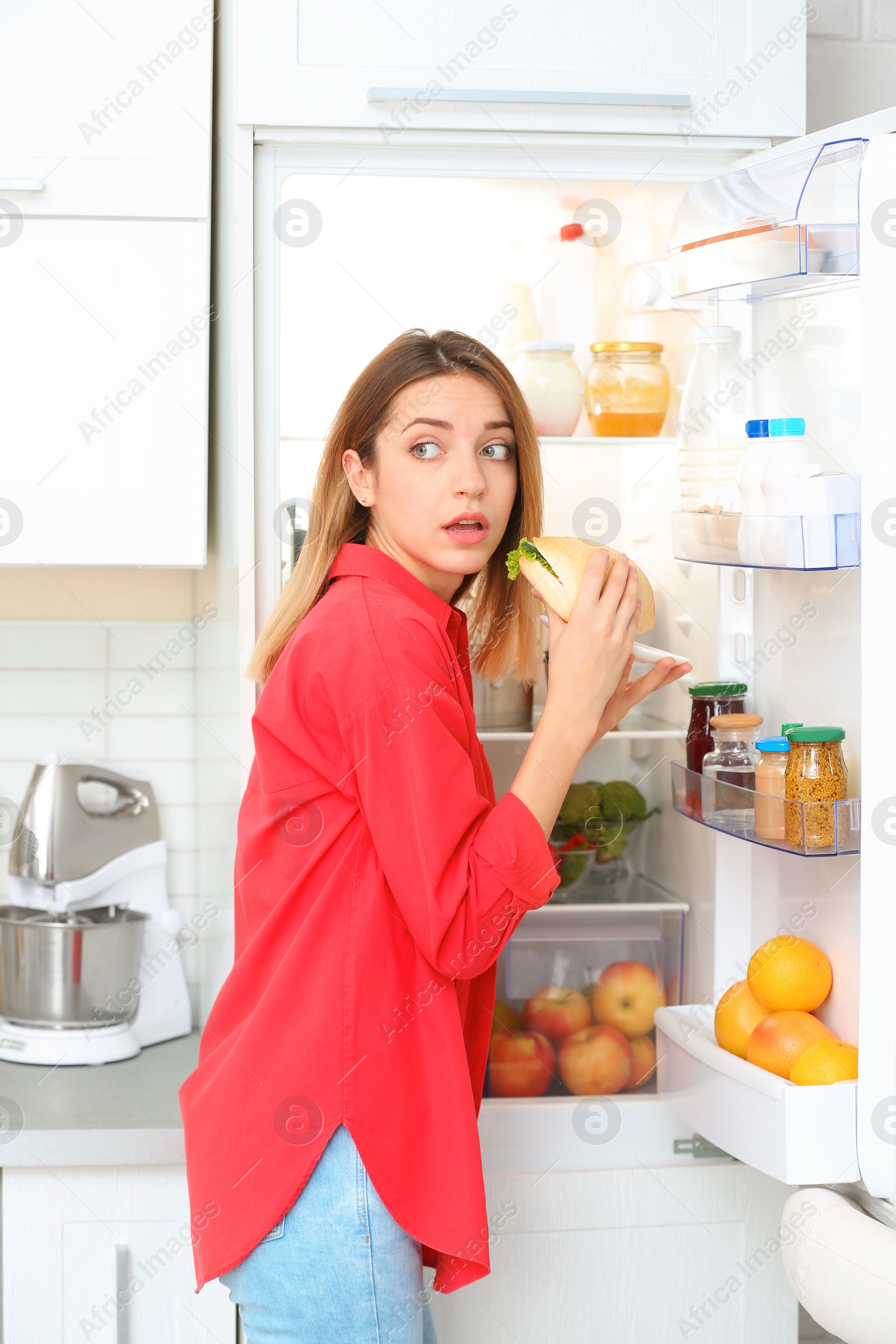 Photo of Emotional young woman eating sandwich near open refrigerator in kitchen. Failed diet