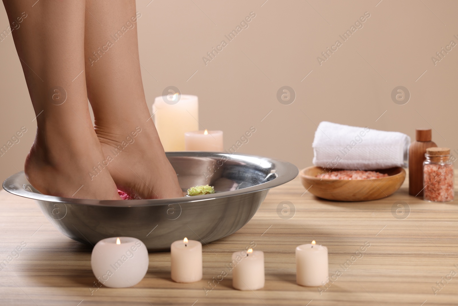 Photo of Woman soaking her feet in bowl with water and flowers on wooden surface, closeup. Pedicure procedure