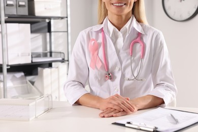 Doctor with stethoscope and pink ribbon at white desk indoors, closeup. Breast cancer awareness