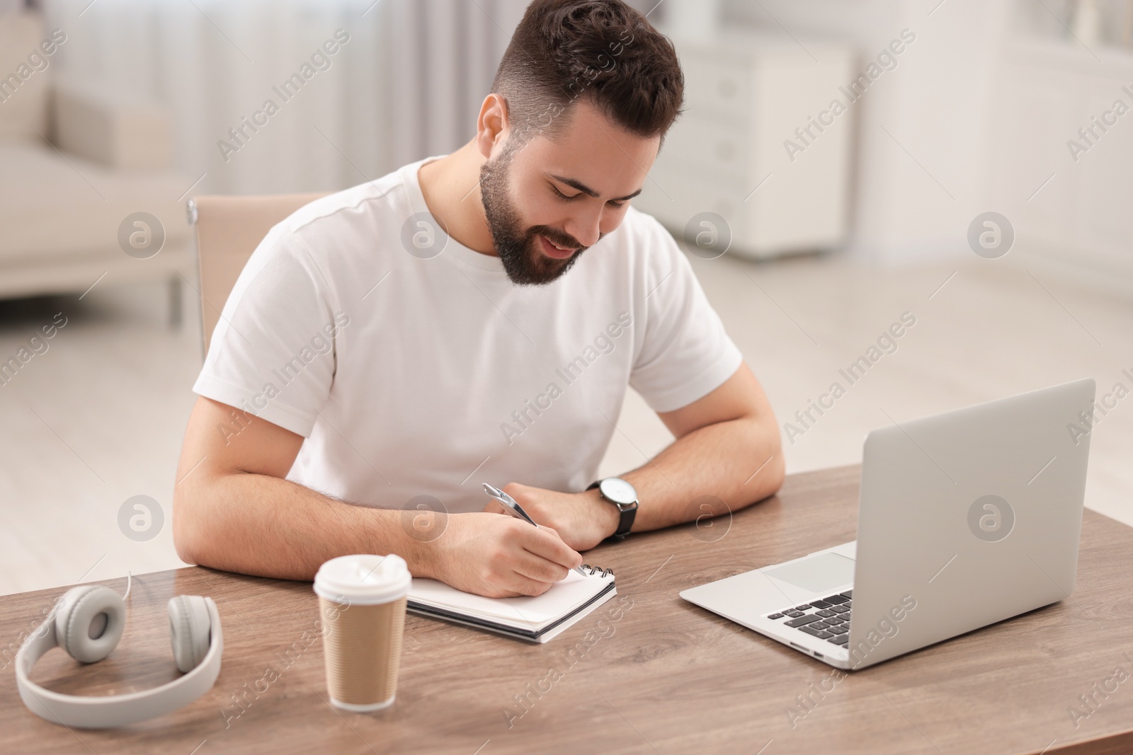 Photo of Young man writing down notes during webinar at table in room