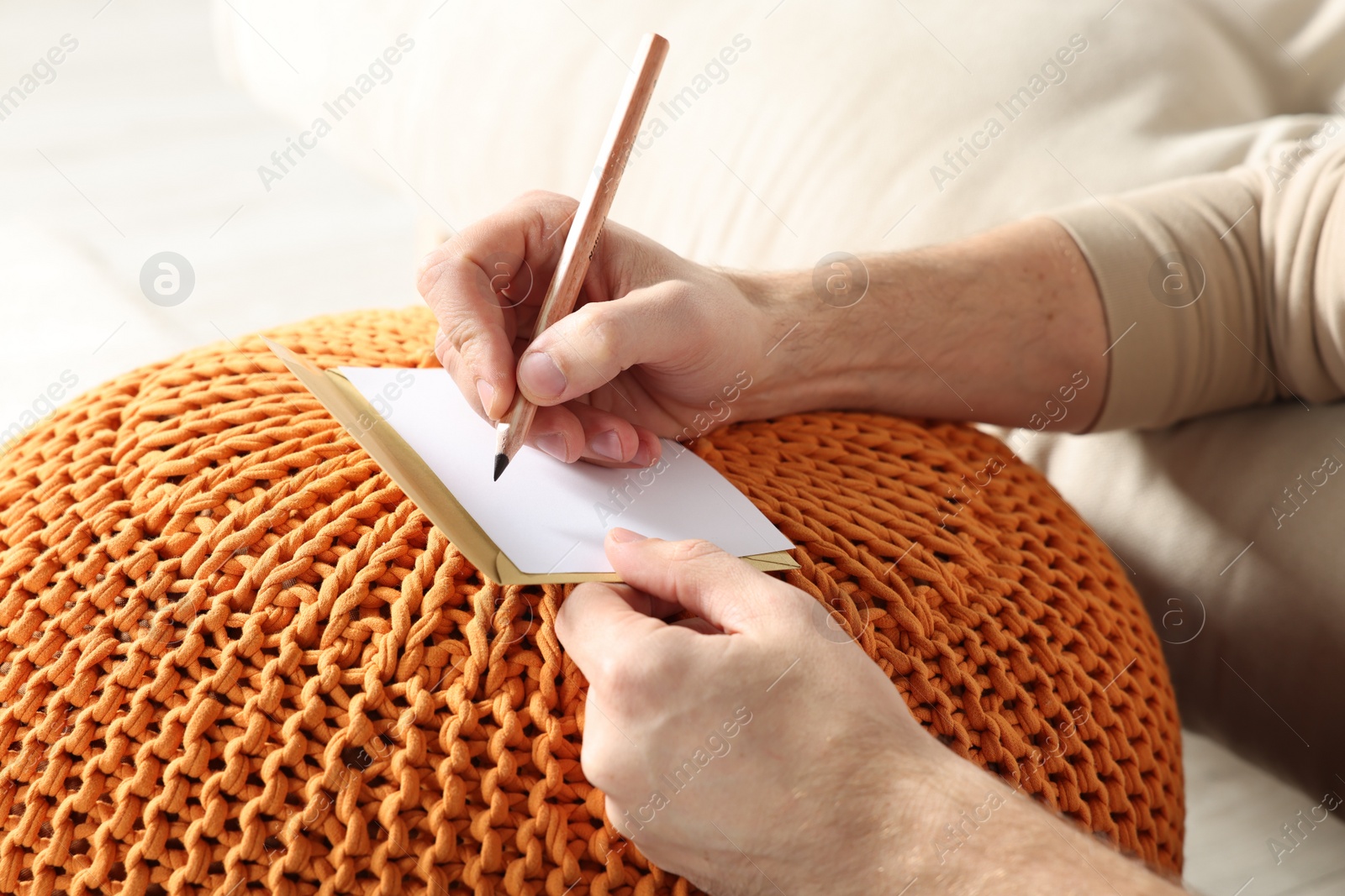 Photo of Man writing message in greeting card on orange
knitted pouf, closeup