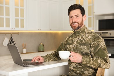 Photo of Happy soldier with cup of drink using laptop at white marble table in kitchen. Military service