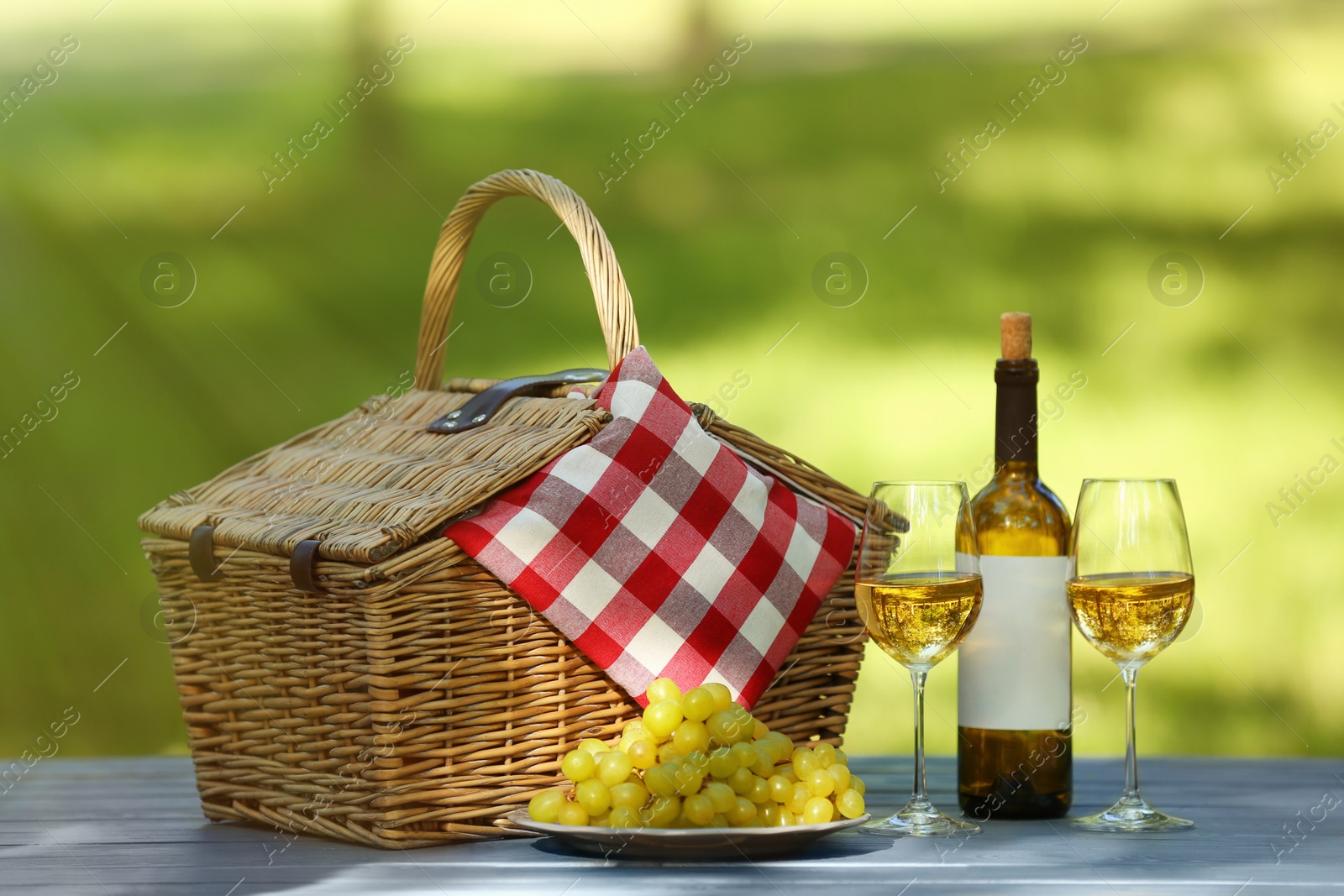 Photo of Wicker basket with blanket, wine and grapes on table in park. Summer picnic