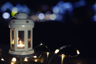 Photo of Lantern and Christmas lights on wooden railing outdoors against blurred background, space for text. Winter night