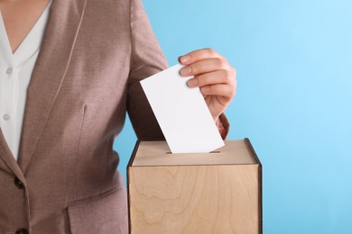 Woman putting her vote into ballot box on light blue background, closeup