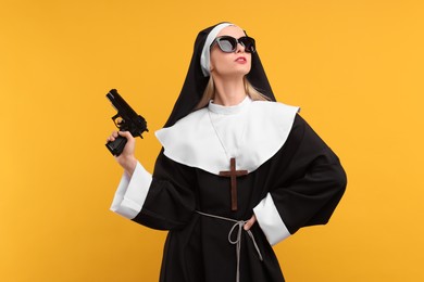 Woman in nun habit and sunglasses holding handgun against orange background