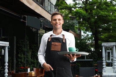 Happy young waiter holding tray with order near cafe