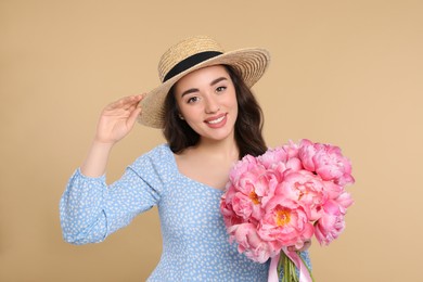 Photo of Beautiful young woman in straw hat with bouquet of pink peonies against beige background