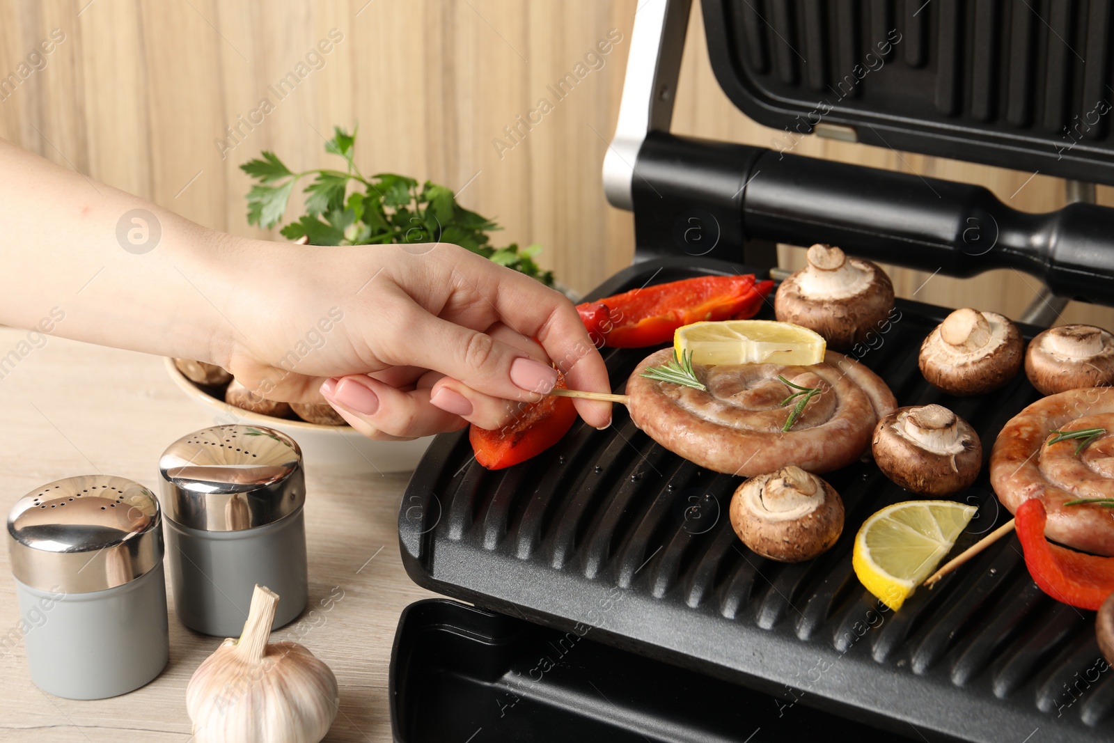 Photo of Woman cooking homemade sausages with mushrooms and bell pepper on electric grill at wooden table, closeup