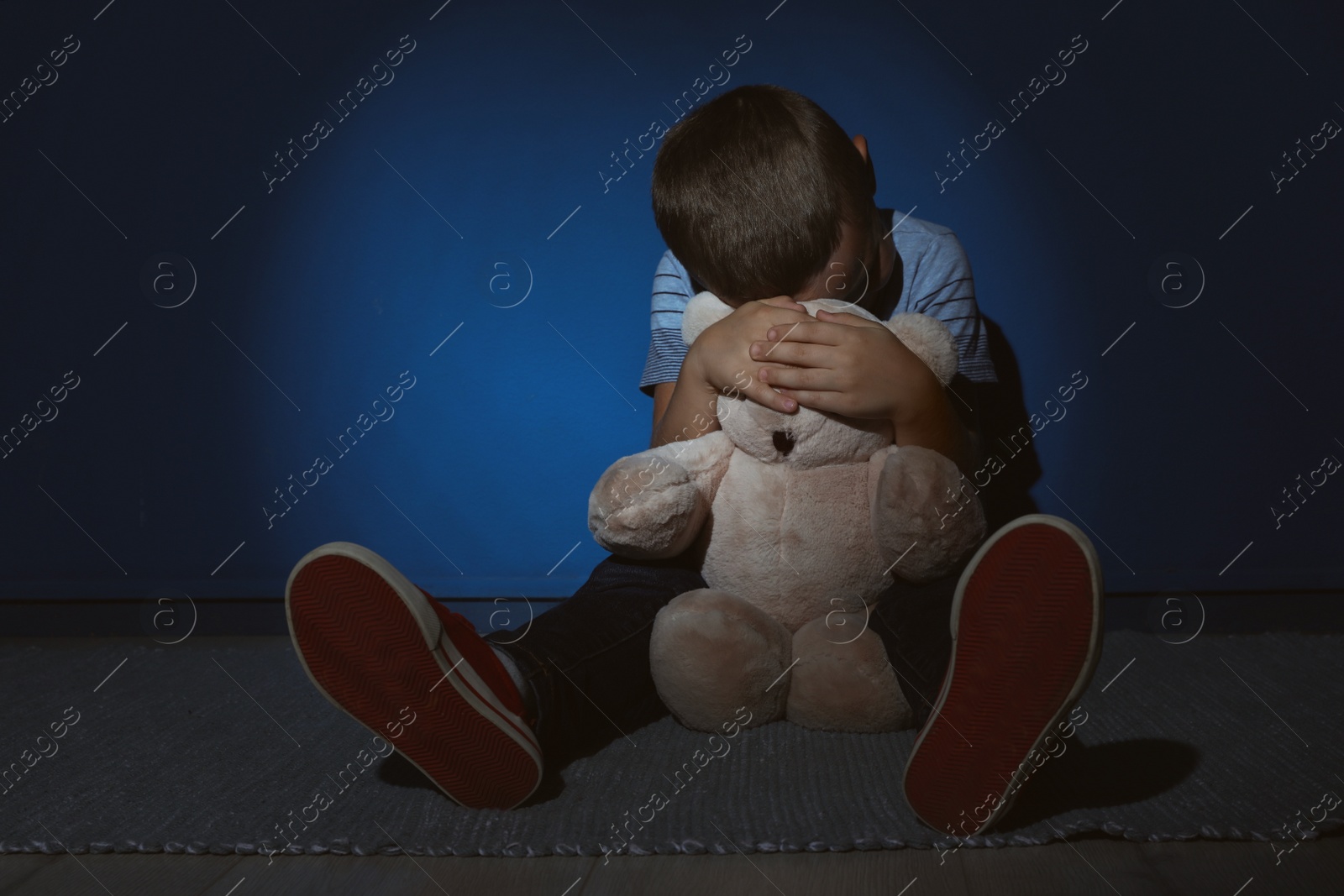 Photo of Sad little boy with teddy bear near blue wall. Domestic violence concept