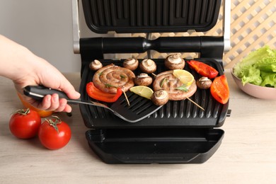 Photo of Woman cooking homemade sausages with bell peppers and mushrooms on electric grill at wooden table, closeup