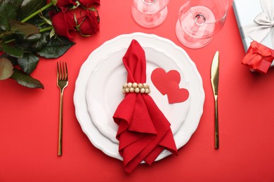 Photo of Place setting with paper hearts, gift boxes and bouquet of roses on red table, flat lay. Romantic dinner