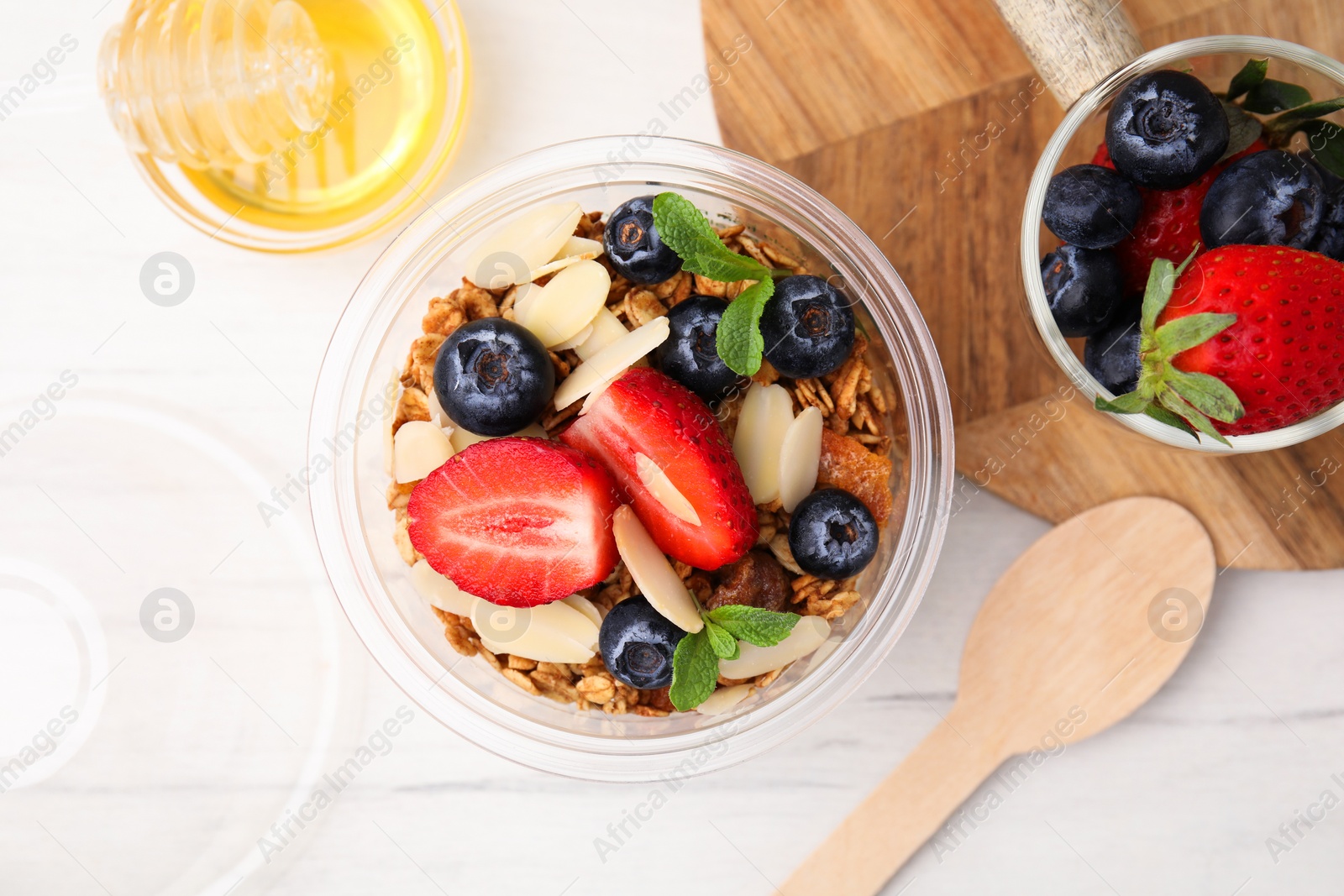 Photo of Tasty granola with berries, almond flakes and mint in plastic cup on white wooden table, flat lay