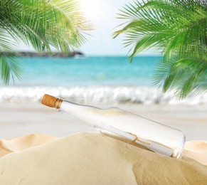Image of Corked glass bottle with rolled paper note on sandy beach with palms near ocean