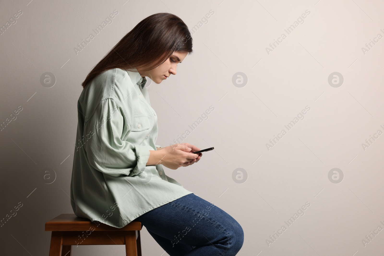 Photo of Young woman with bad posture using smartphone while sitting on stool against grey background. Space for text