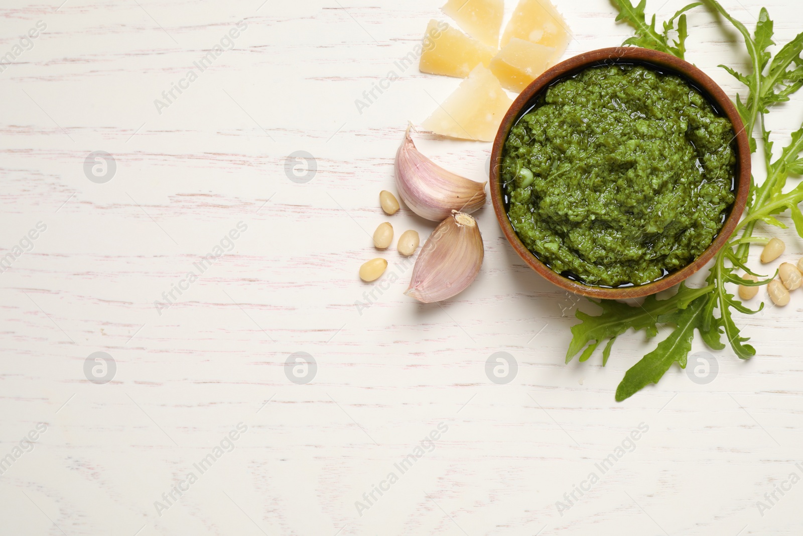 Photo of Bowl of tasty arugula pesto and ingredients on white wooden table, flat lay. Space for text