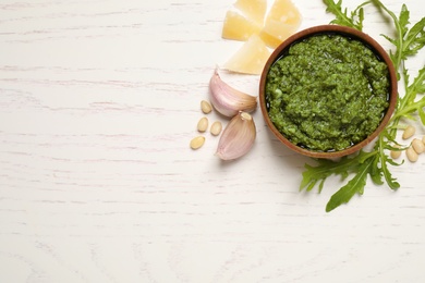 Photo of Bowl of tasty arugula pesto and ingredients on white wooden table, flat lay. Space for text