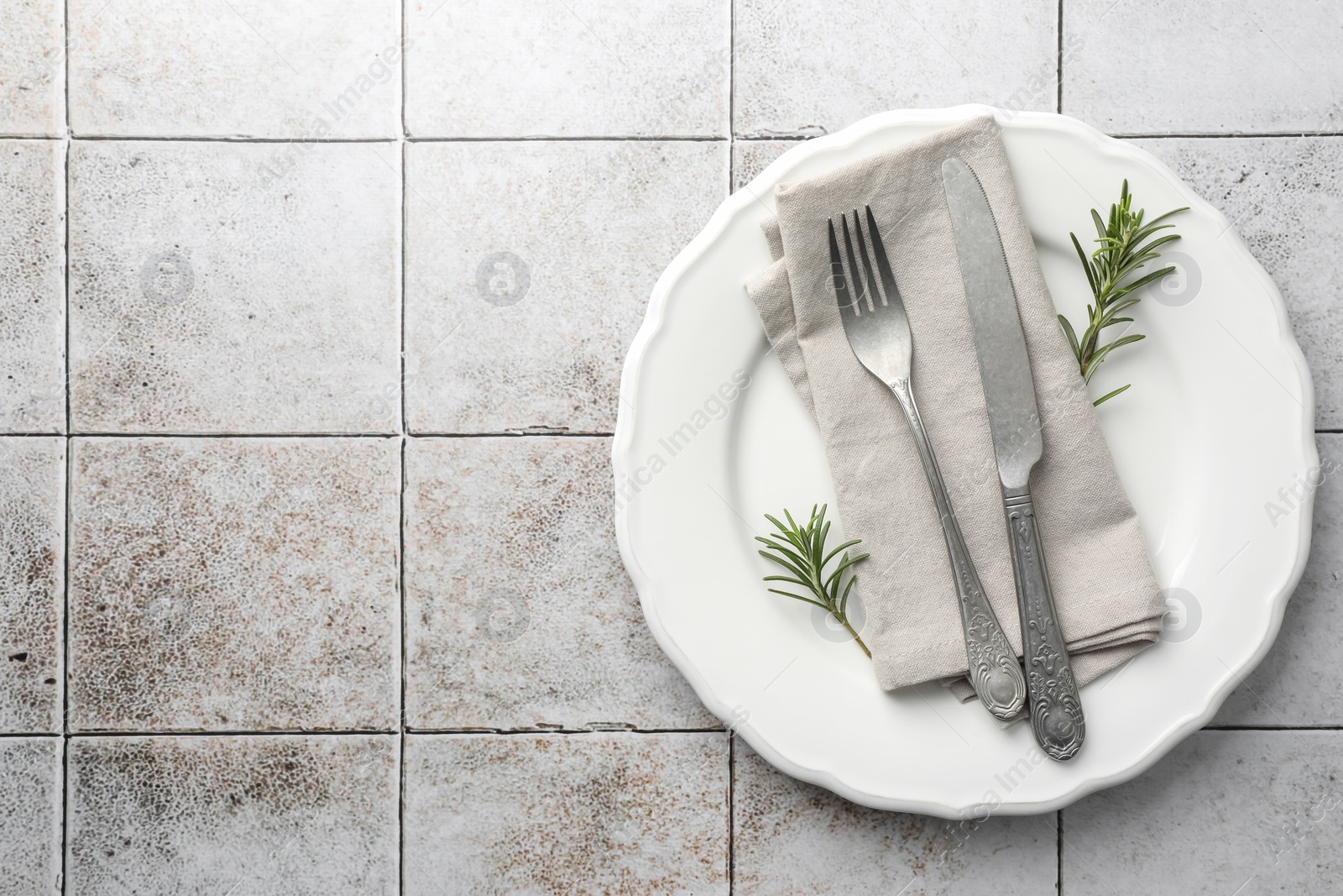 Photo of Stylish setting with cutlery, napkin, rosemary and plate on light tiled table, top view. Space for text