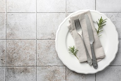 Stylish setting with cutlery, napkin, rosemary and plate on light tiled table, top view. Space for text