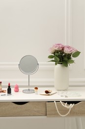 Photo of Mirror, cosmetic products, jewelry and vase with pink roses on white dressing table in makeup room
